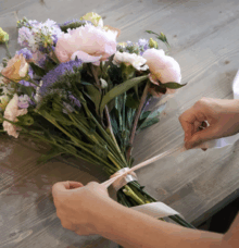Händer binder ett band runt en bukett med olika blommor på ett träbord.